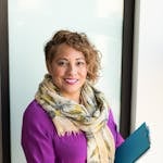 Smiling woman in a purple dress and scarf holding a tablet indoors.